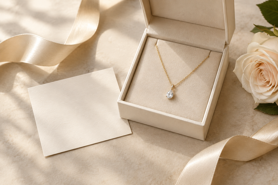 Illustrative necklace gift in an ivory jewelry box beside a blank note card, satin ribbon, and a pale rose.