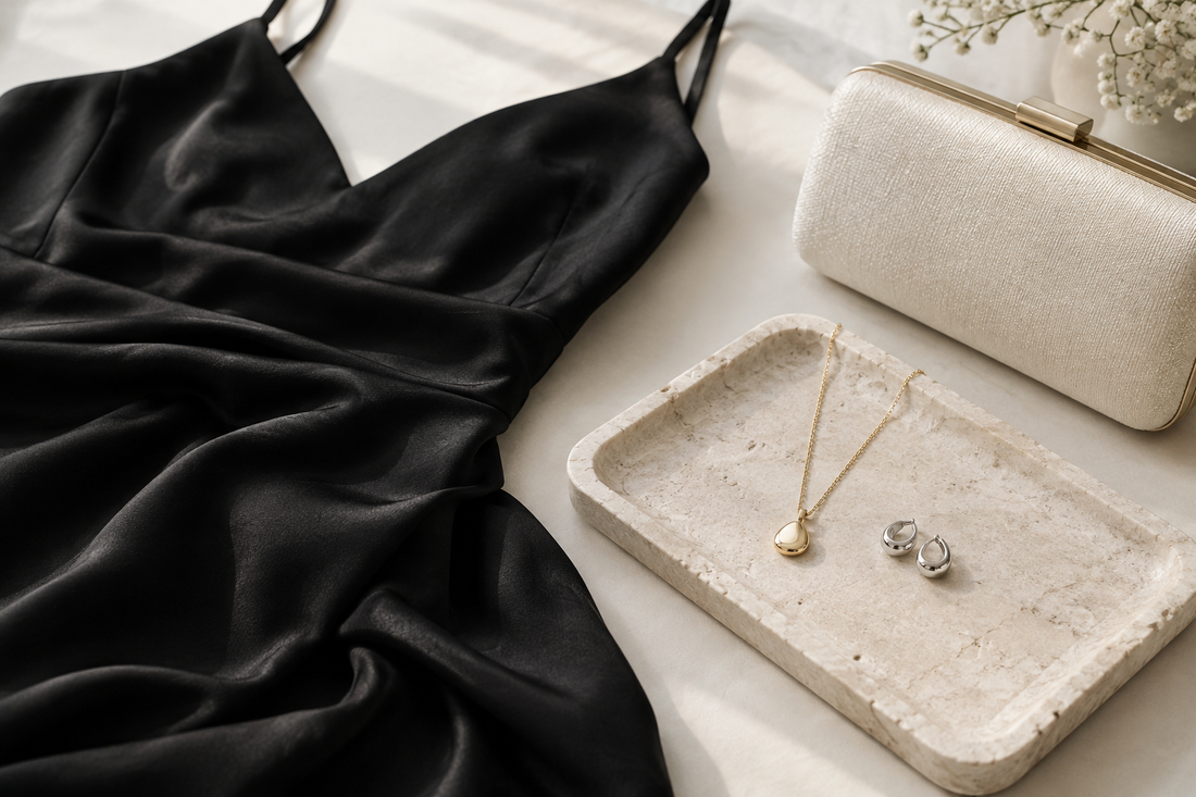 Illustrative gold-tone necklace and silver-tone earrings beside black dress fabric and a cream clutch.