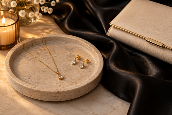 Illustrative gold-tone necklace and earrings beside black satin fabric and a cream clutch in warm evening light.