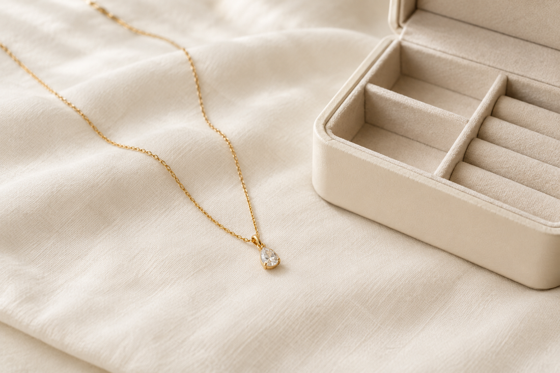 Illustrative gold-tone pendant necklace on a soft ivory cloth beside an open cream jewelry box.
