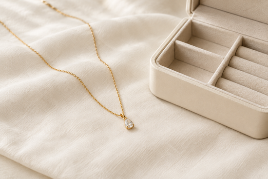 Illustrative gold-tone pendant necklace on a soft ivory cloth beside an open cream jewelry box.