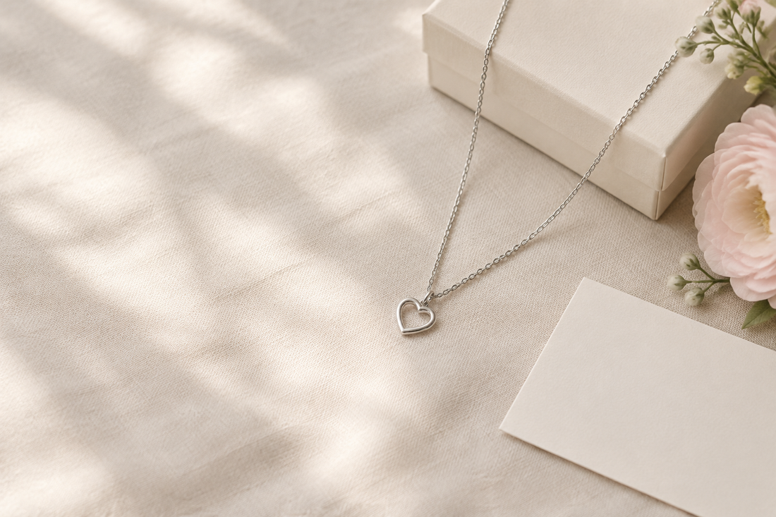 Illustrative silver-tone open-heart pendant necklace on ivory linen beside a cream gift box and blank note card.