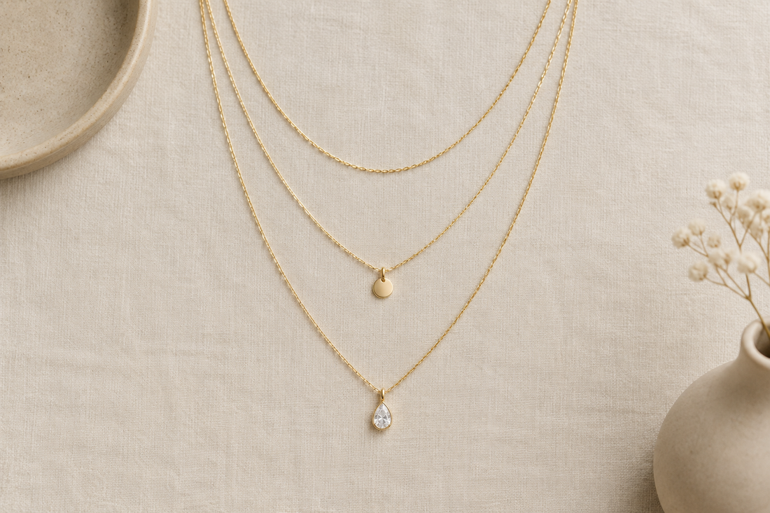 Illustrative arrangement of three gold-tone necklaces at different lengths on ivory linen, including round and drop-shaped pendants.