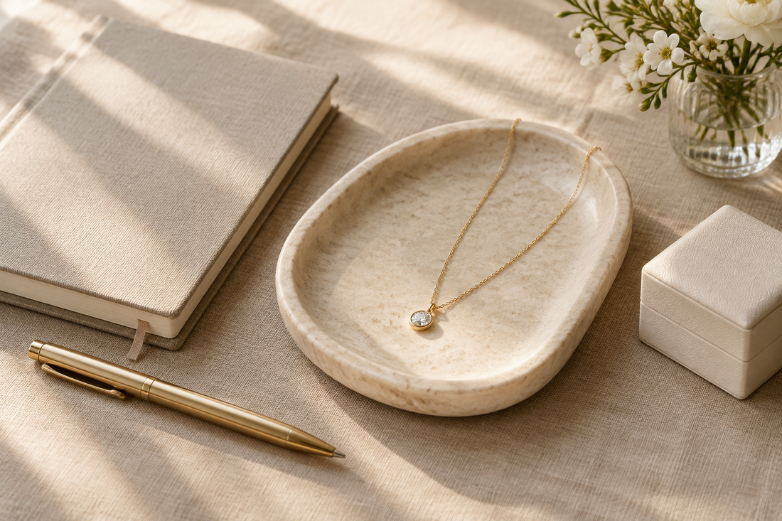 Illustrative gold-tone pendant necklace beside a journal, pen, and ivory jewelry box in soft morning light.