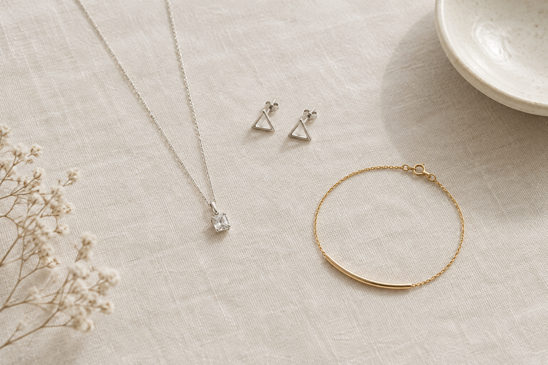 Illustrative minimalist jewelry arrangement with a pendant necklace, triangular studs, and a slim bracelet on ivory linen.