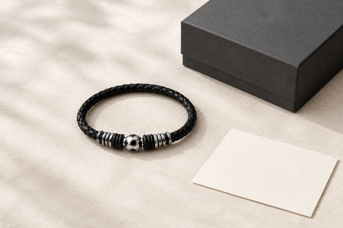 Black braided soccer bracelet with a silver-tone ball centerpiece arranged beside a charcoal gift box and blank note card.