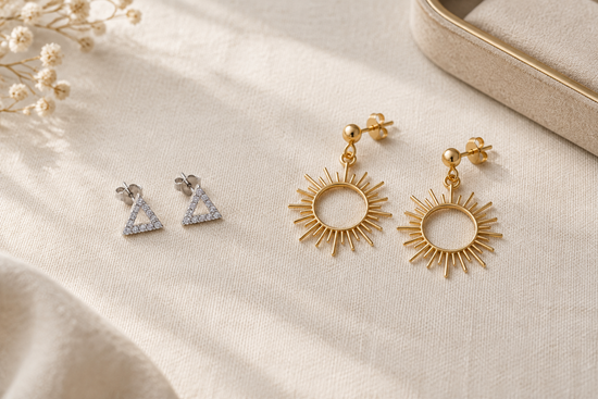 Illustrative comparison of silver-tone triangular studs and gold-tone dangling sunburst earrings on ivory linen.