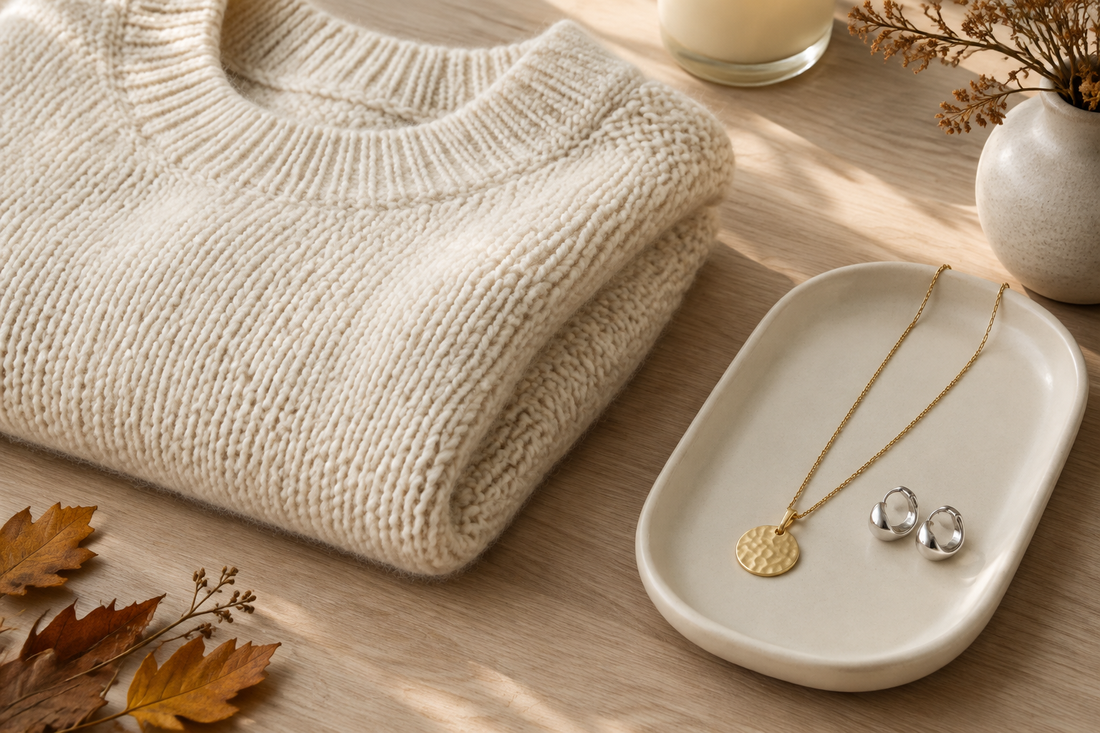 Illustrative cream knit sweater beside a gold-tone pendant necklace and silver-tone earrings on an ivory tray.