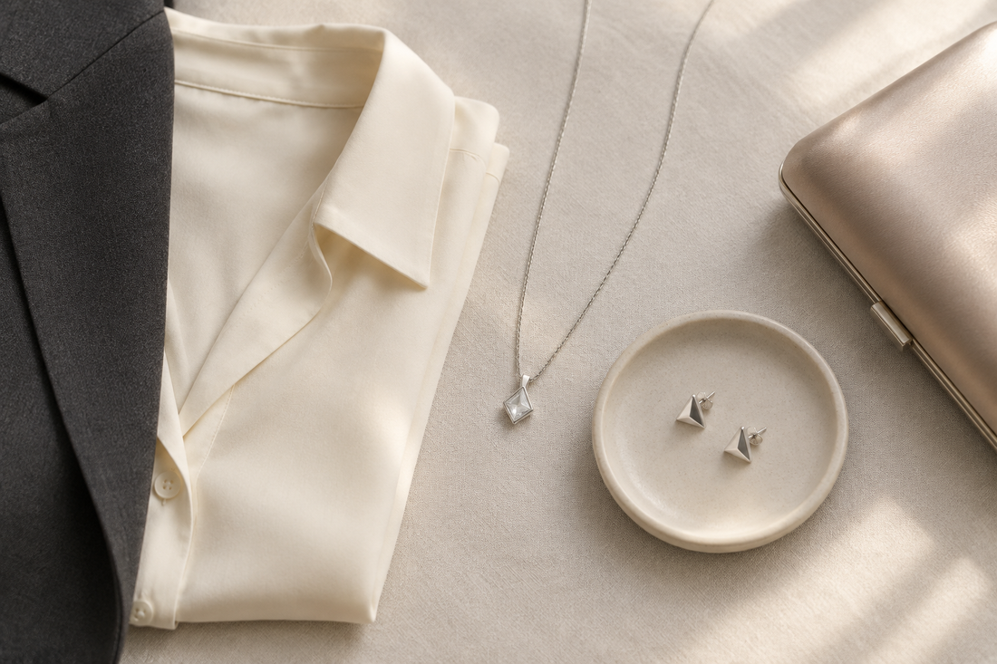 Illustrative pendant necklace and triangular studs beside a cream blouse, charcoal blazer, and satin clutch.